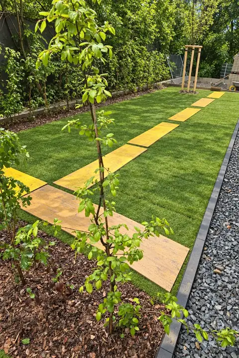 Frisch verlegter Rollrasen mit jungem Baum, darauf gelbe Platten als Laufweg, rechts dunkler Splitt