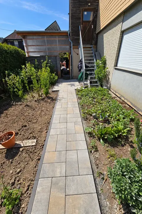 Gerader Plattenweg zwischen frisch angelegten Beeten am Wohnhaus, hinten eine Außentreppe