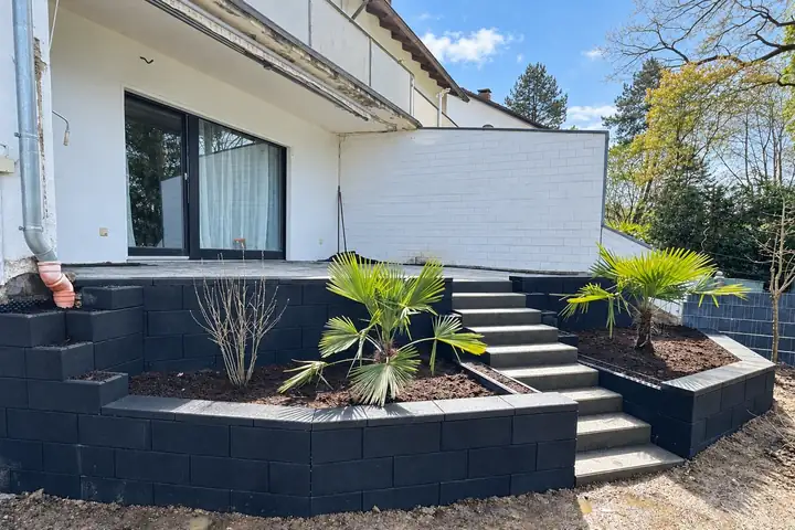 Hangabfangung aus anthrazitfarbenen Blocksteinen mit Treppe und Palmen vor einem weißen Wohnhaus