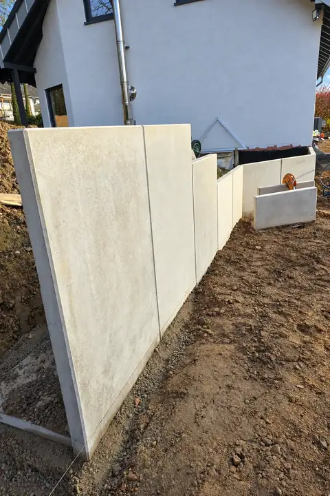 Helle L-Steine als gestaffelte Stützwand auf vorbereitetem Boden an einem weißen Wohnhaus