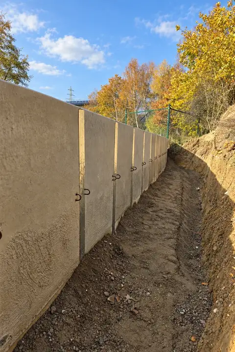 Lange Stützmauer aus L-Steinen entlang eines Erdgrabens, dahinter Herbstbäume und Zaun