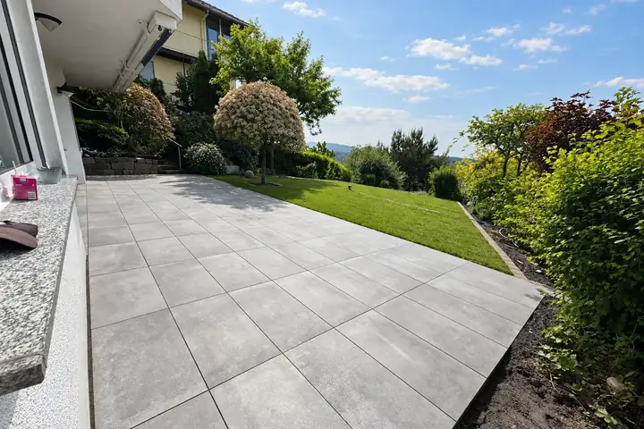 Terrasse aus großformatigen grauen Platten am Haus, daneben Rasen und Gehölzrand