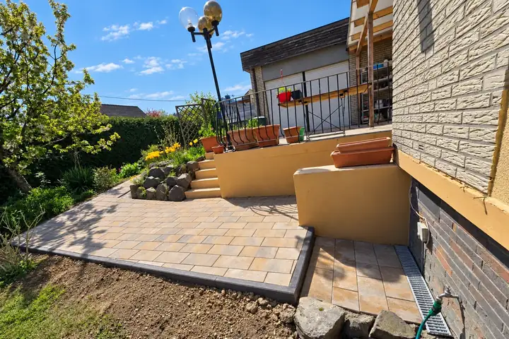 Terrasse aus sandfarbenem Rechteckpflaster mit dunkler Einfassung, Treppe und Steinbeet am Wohnhaus
